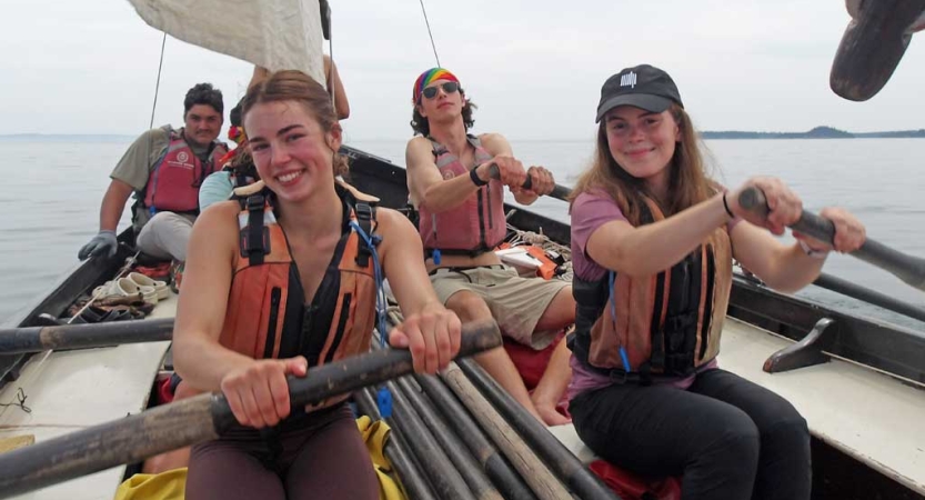 Four students wearing life jackets navigate a sailboat using oars. 